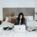 Woman sitting on a bed working on a laptop earning $99,000 a year.